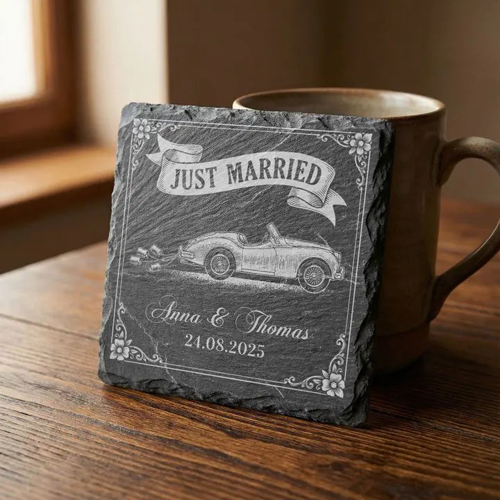 Schiefer Untersetzer mit Hochzeitsauto-Gravur Just Married lehnt an Keramikbecher auf Holztisch