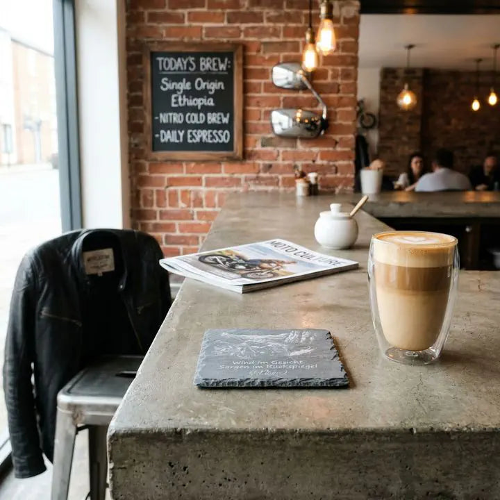 Schiefer Untersetzer mit Biker-Motiv und Spruch Wind im Gesicht neben Cappuccino im Café